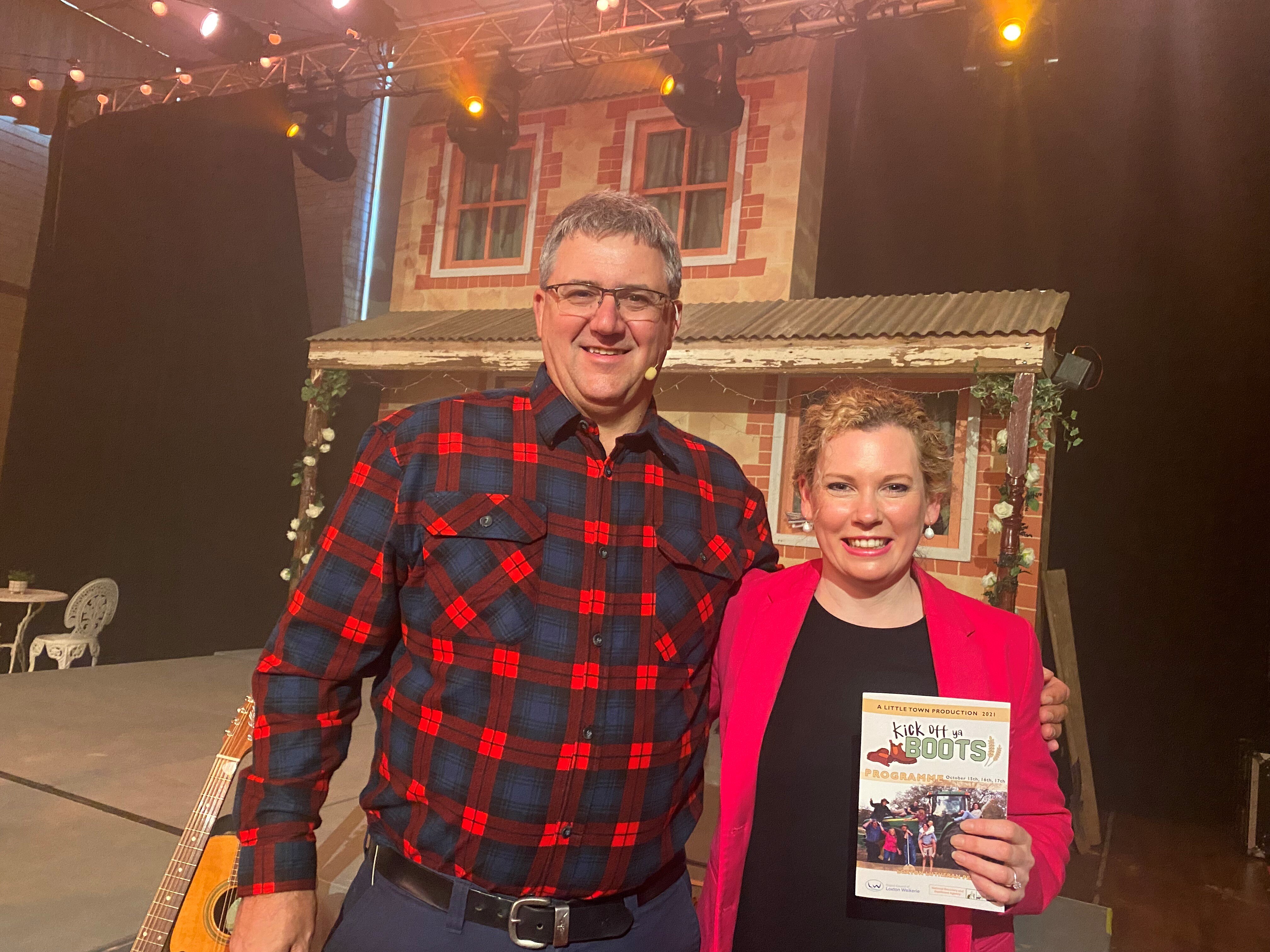 A smiling white man in a checked shirt has his arm around a smiling blonde white woman in black and a pink blazer in a theatre. 