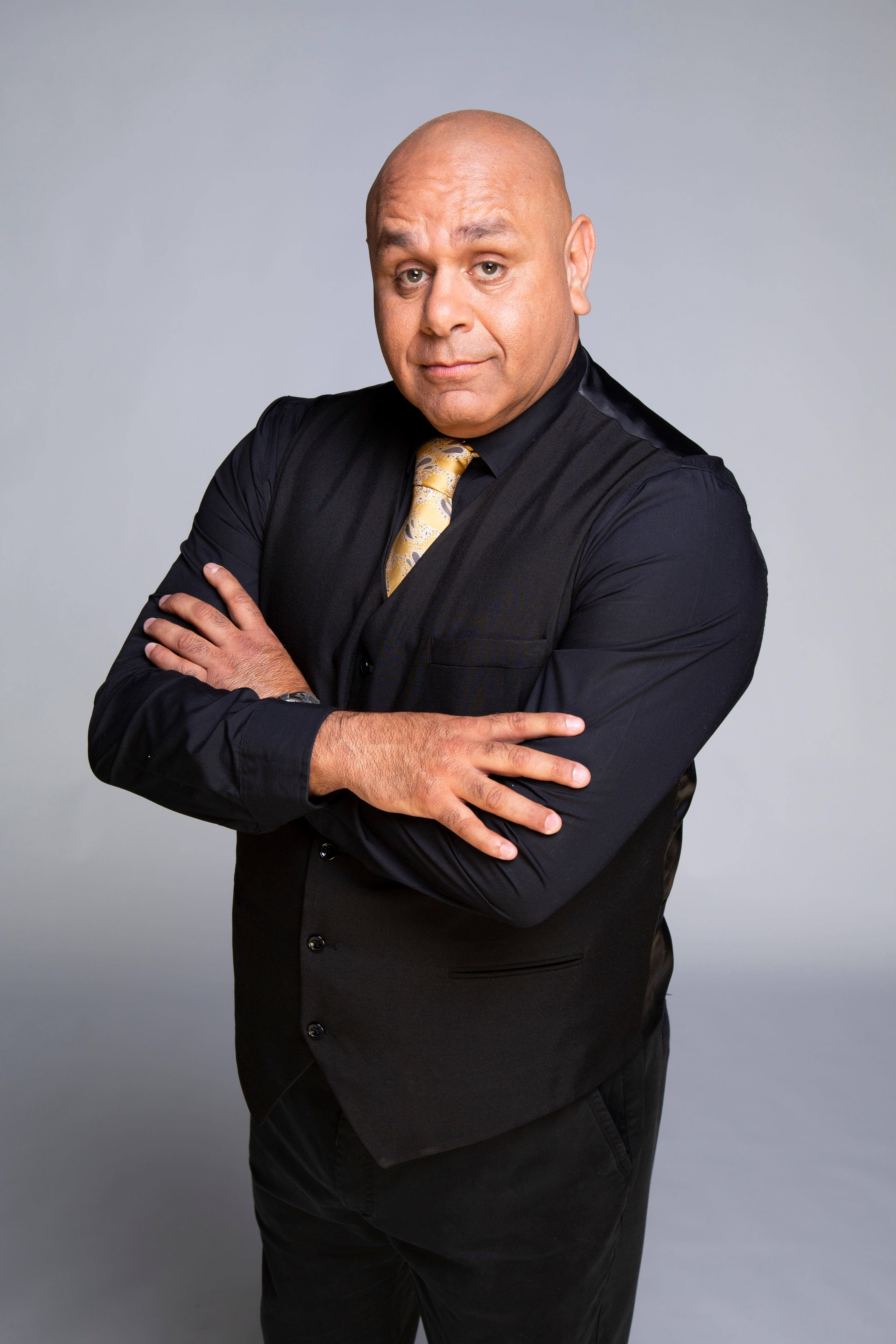 A man in a black suit and yellow tie looks at the camera with a slightly raised eyebrow and folded arms