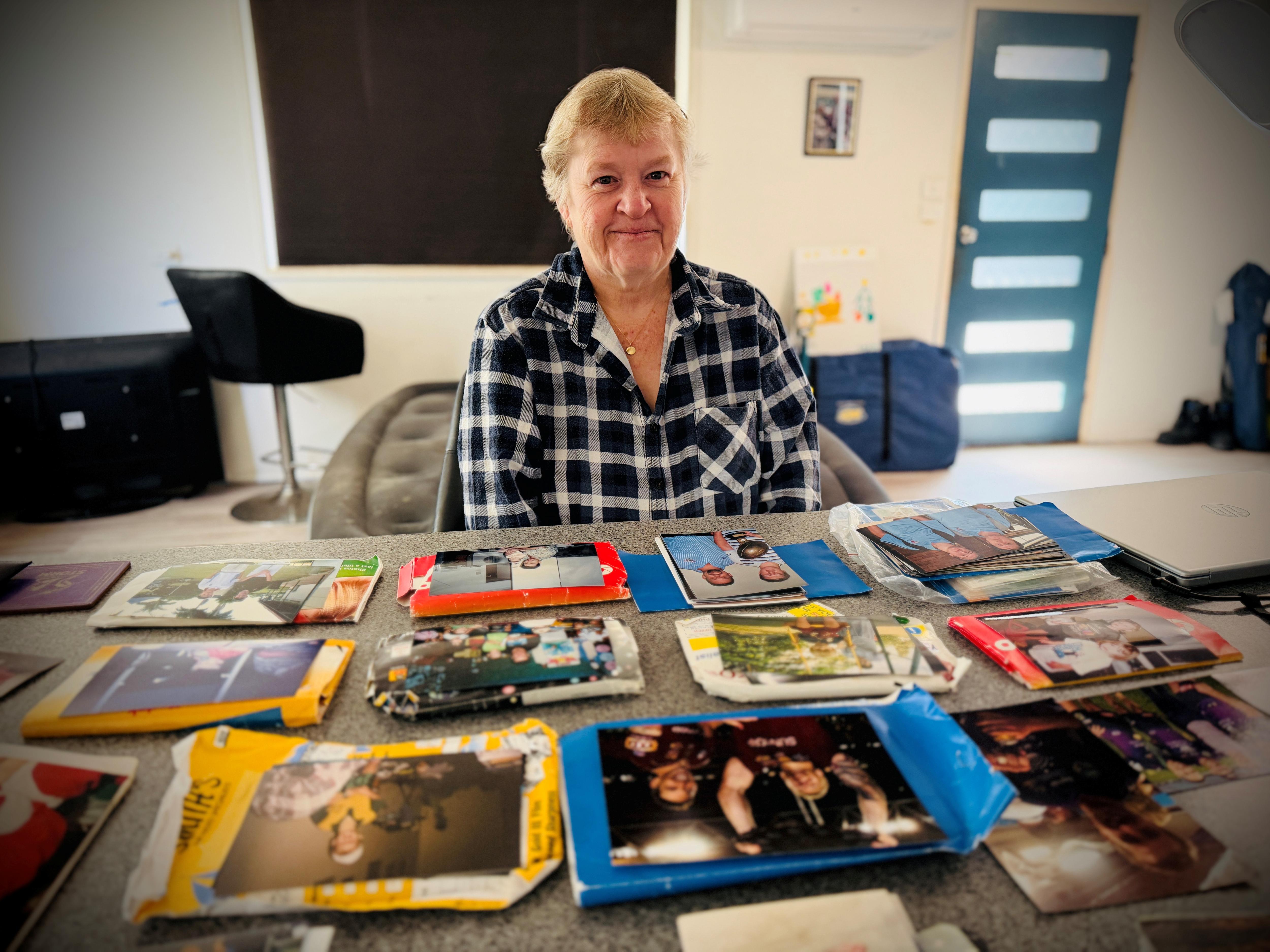Debbie smiles as she sits in front of a benchtop of old photos, she has short blonde hair, blue front door, tv behind.