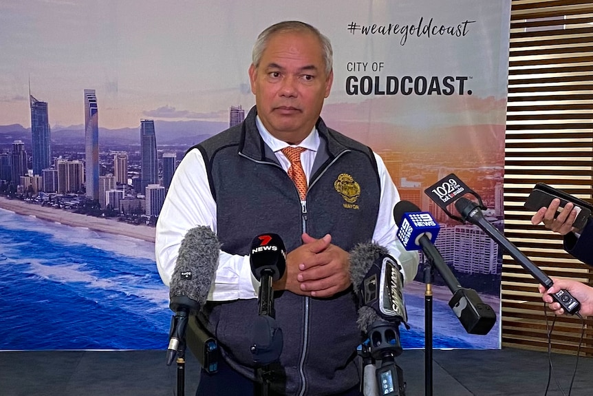  a balding man speaks into microphones in front of a banner of the Gold Coast