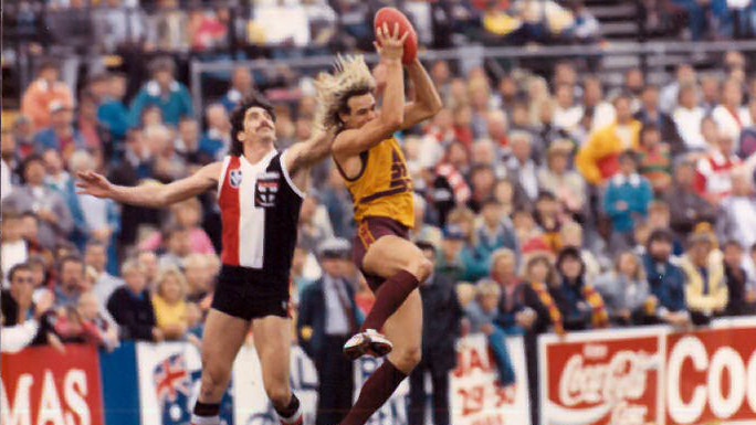 Man with blonde hair kicking in VFL Brisbane Bears uniform on a field in front of an crowd.