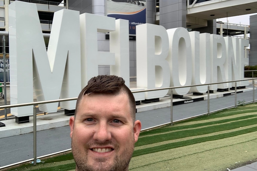 A smiling man stands in front of a sign reading "Melbourne" at Melbourne Airport.