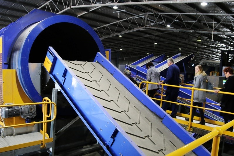 Several people visiting a recycling plant Canberra walking past machinery and a conveyor belt used to sort recyclable waste.