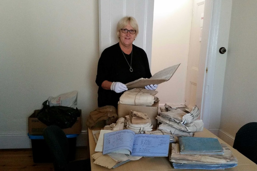 Di Hausler wearing white gloves and standing behind a table loaded with stacks of old pages and books.