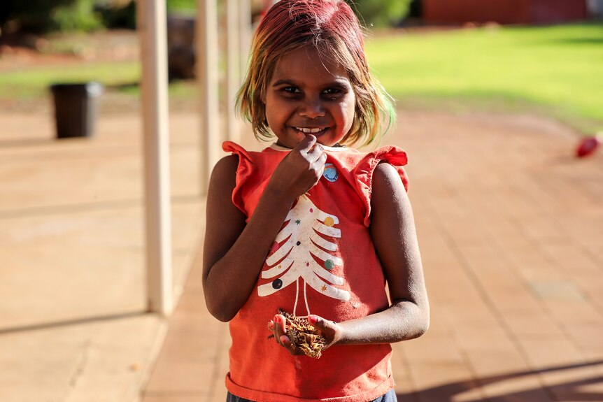 Young Aboriginal girl with died hair and red shirt smiles holding an brightly coloured thorny devil insect