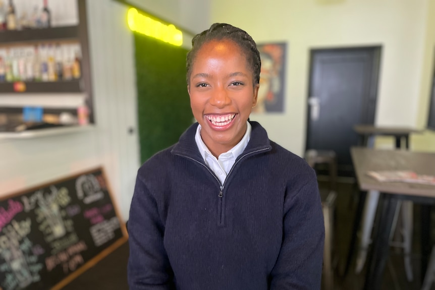 A woman with a big smile sits in a room with a table and blurred sign in the background