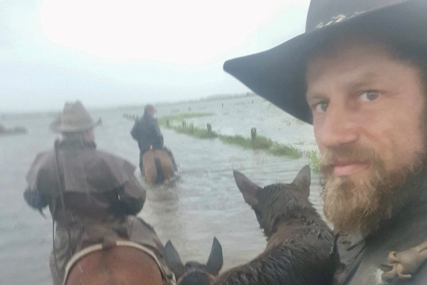 A bearded man in a hat atop a horse in floodwaters looks at the camera. Two other men on horses are ahead.