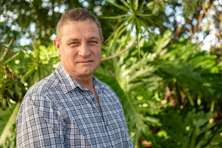 Man wearing a blue checkered shirt standing in front of a tree.