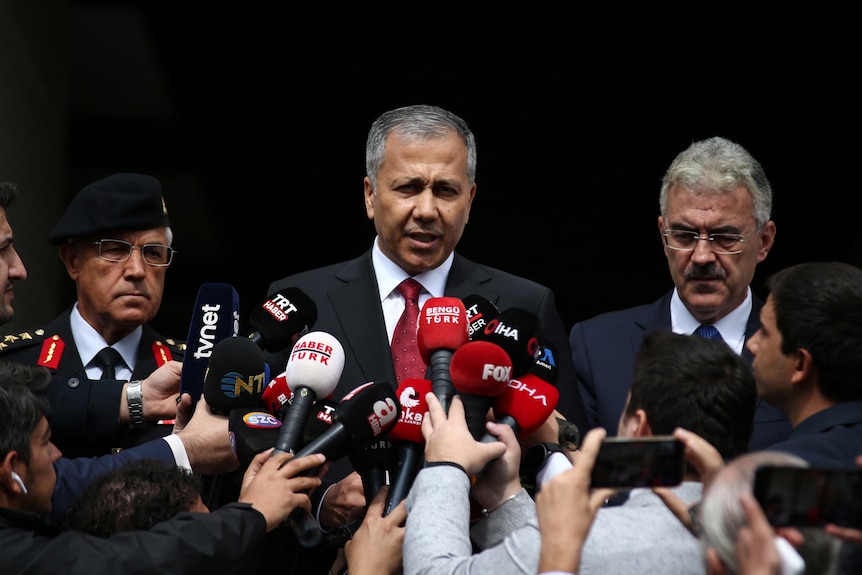 Ali Yerlikaya speaking in front of several media microphones. 