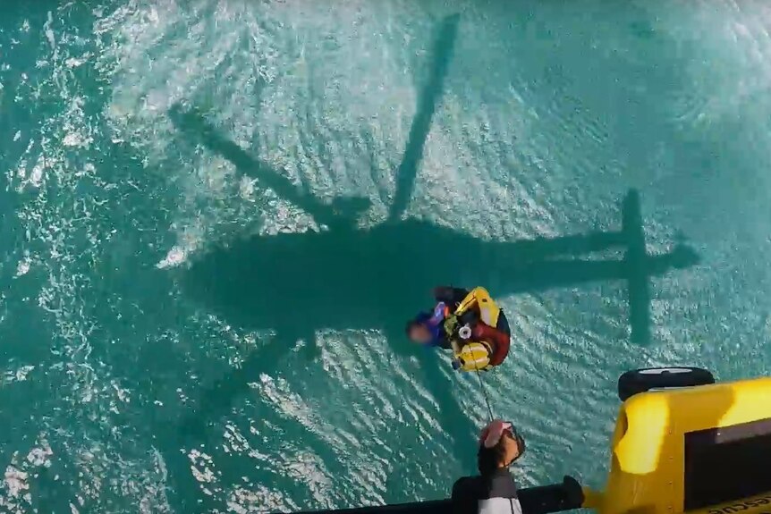 Birds-eye view of a person below a helicopter on a rope holding a second person, above the ocean