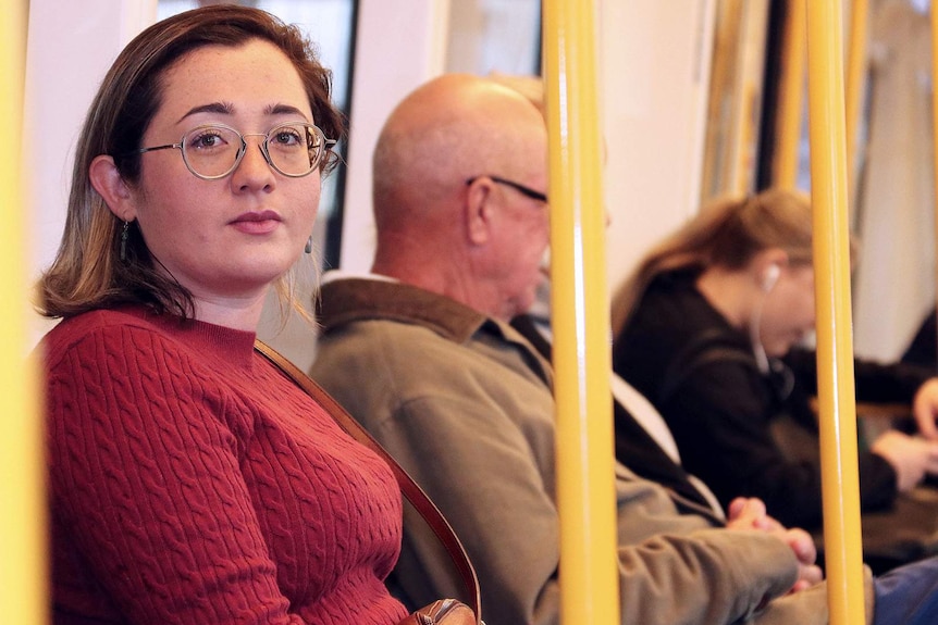A mid shot of a woman wearing a red jumper and spectacles sitting on a train and looking at the camera.