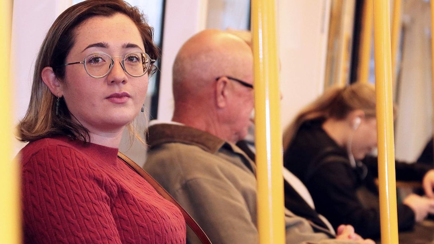 A mid shot of a woman wearing a red jumper and spectacles sitting on a train and looking at the camera.
