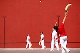 A group of men. Some stand in front of a red wall with the curved basket gloves used in pelota. One man is throwing a ball.