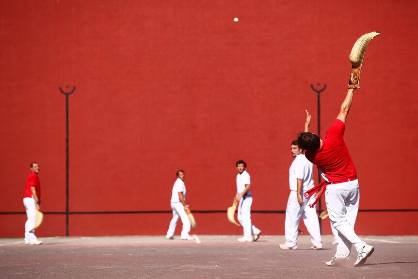 A group of men. Some stand in front of a red wall with the curved basket gloves used in pelota. One man is throwing a ball.