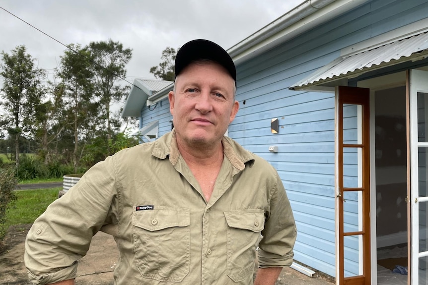 Man wearing khaki button up shirt and cap, hands on his hips, unsmiling, standing in front of a blue weatherboard house