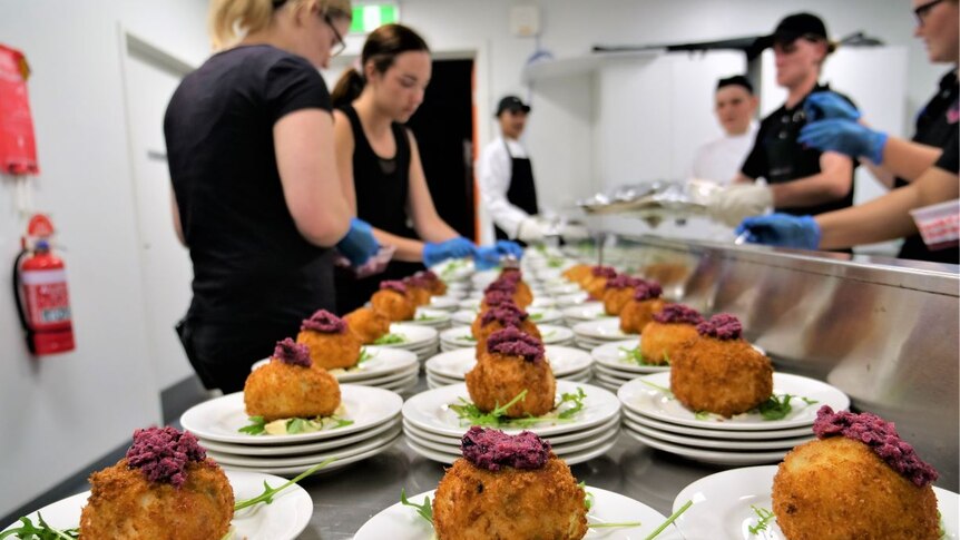 Plates of arancini and garnishes lined up on an industrial kitchen bench with students wearing blue gloves in background.