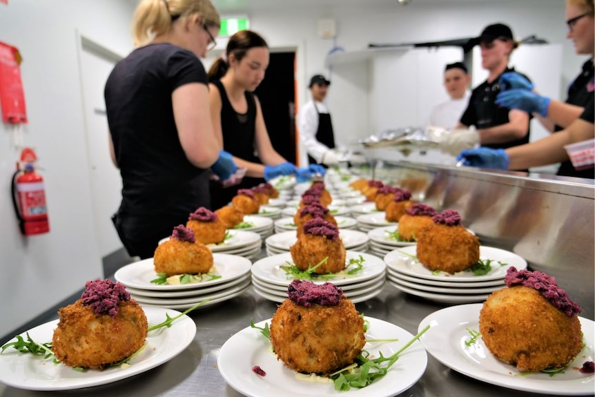 Plates of arancini and garnishes lined up on an industrial kitchen bench with students wearing blue gloves in background.