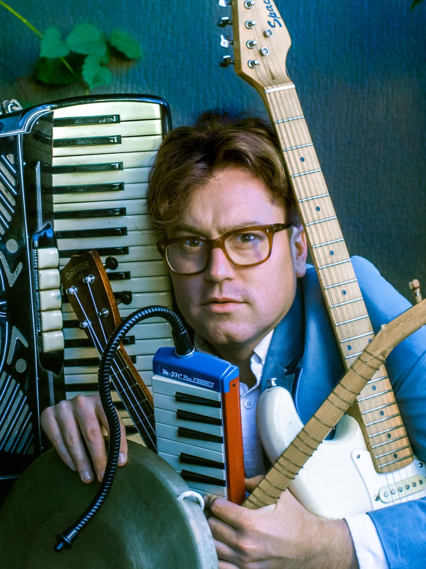 Joff Bush poses with instruments stacked around his head & body including an accordion, melodica, ukulele, electric guitar &gong