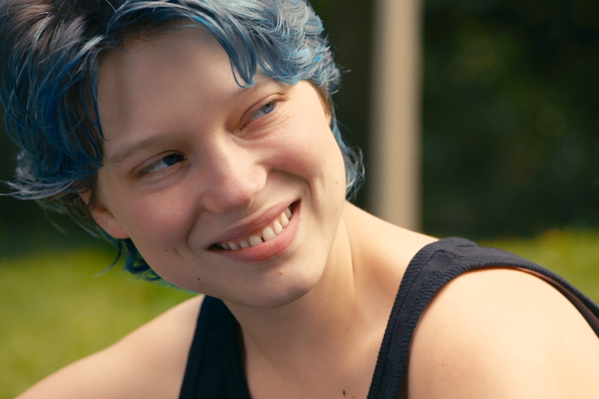 A young woman smiles broadly at someone out of frame. She has cropped hair, dyed blue, and is wearing a black singlet. 