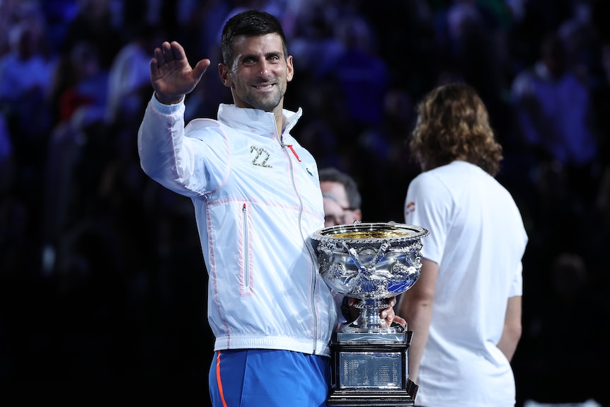 Novak Djokovic waves with his right hand as he holds the Australian Open trophy with his left.