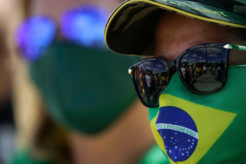 A close up image shows the face of a person wearing a Brazilian flag for a face mask, dark sunglasses and an army baseball cap.