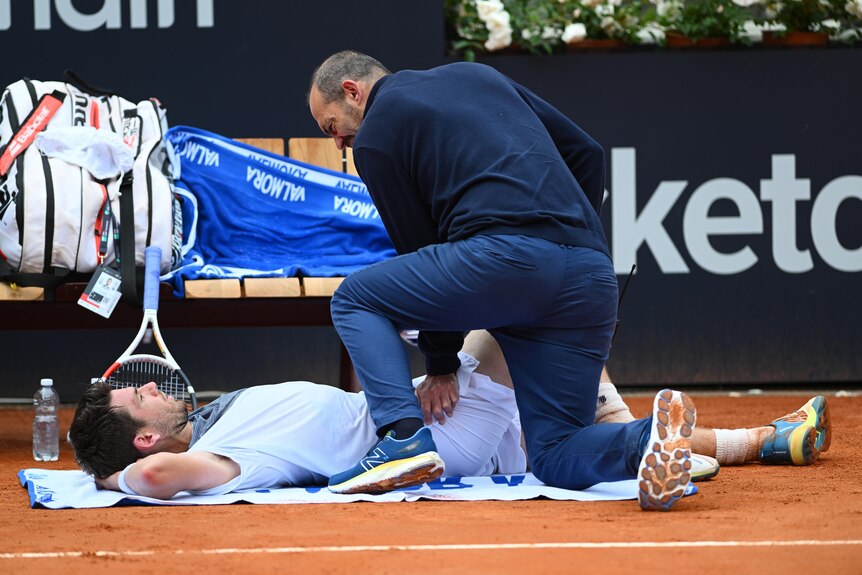 A tennis player lies flat on his back on the red clay court as a trainer works on him during a medical timeout.