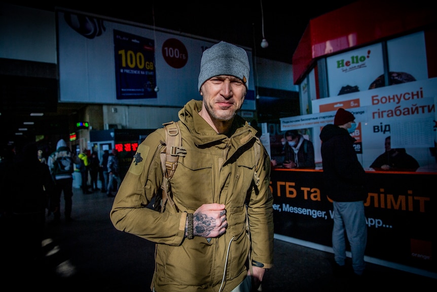A man wearing a coat and beanie with a tattoo on his arm smiles at the canera.