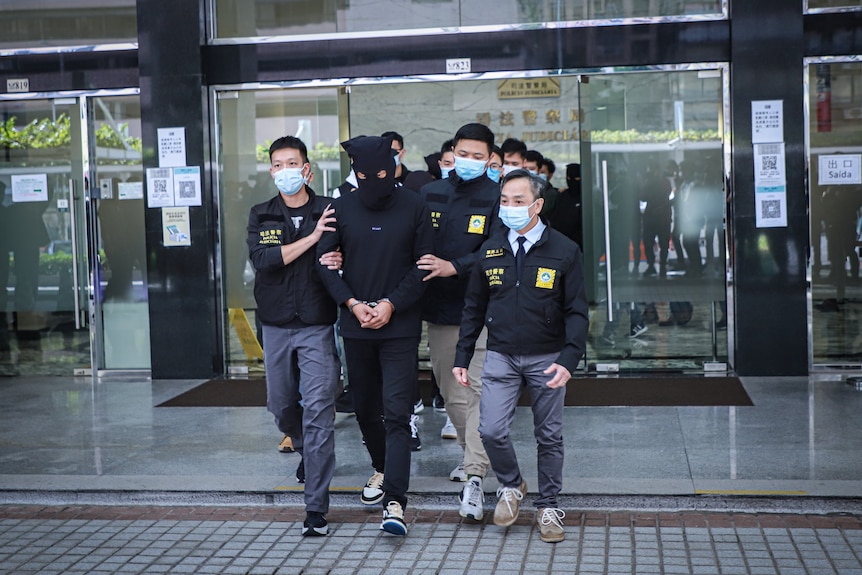 Un grupo de policías camina con un hombre cubierto con una máscara negra. 