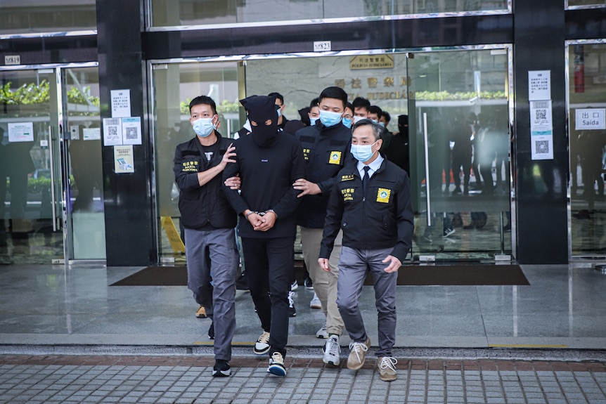 A group of police walk with a man covered in a black mask. 