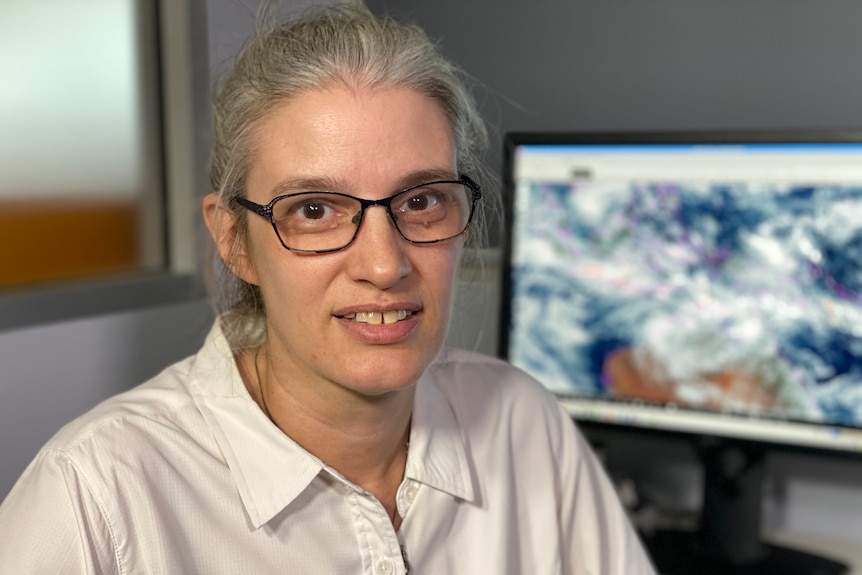 A woman with black-rimmed glasses and grey hair smiles at the camera, with a forecast map in the backgroun