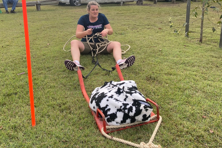 A woman sits on the ground with a rope leading to a metal sled with a black and white sandbag on it.