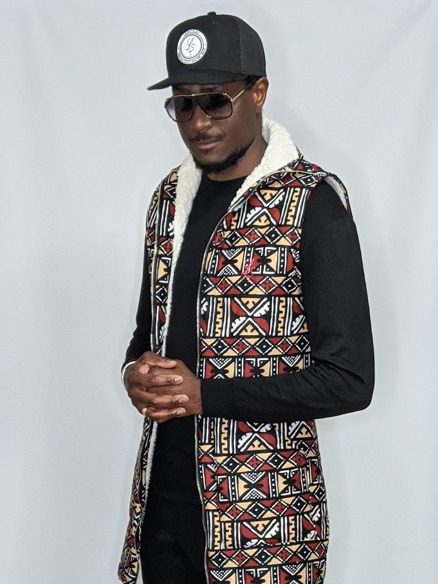 A very tall and slim black man wearing a baseball cap, sunglasses and black clothing with a sleeveless colourful jacket