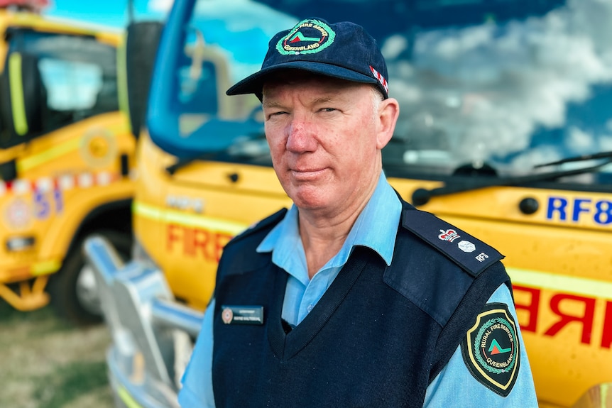 A man in a fire rescue uniform stands in front of two yellow rural fire trucks