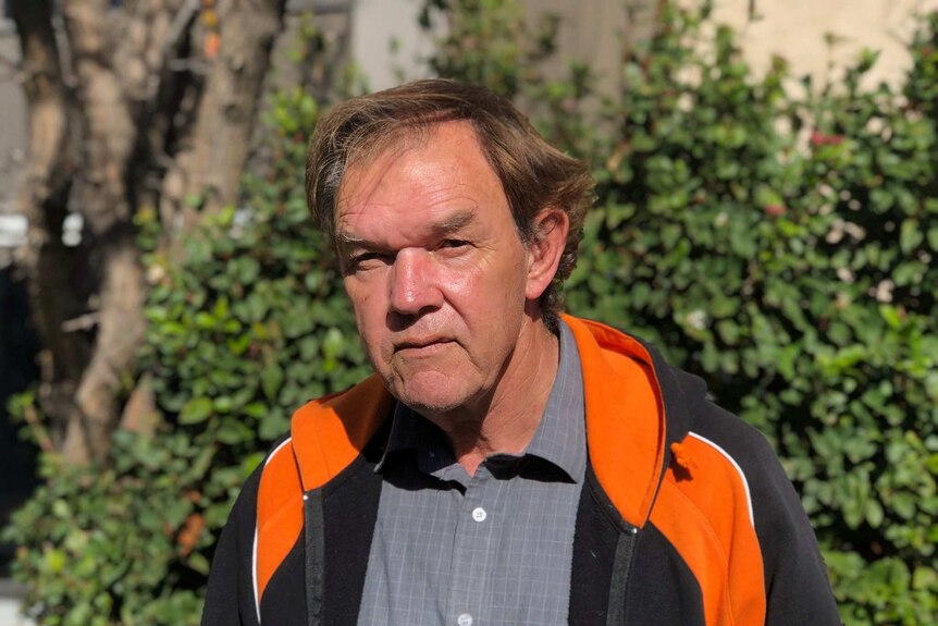 A man stands in front of shrub, he is wearing a black and orange jacket.
