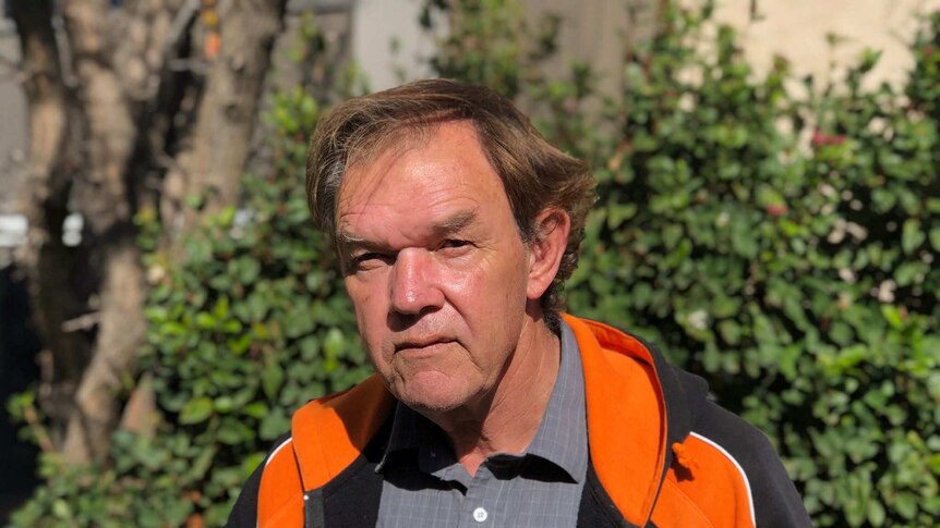 A man stands in front of shrub, he's wearing a black and orange jacket.