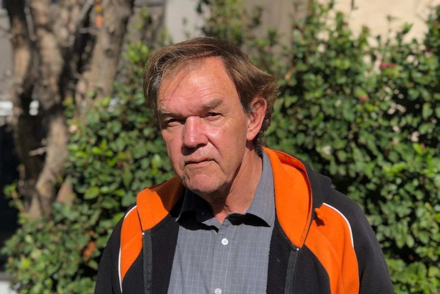 A man stands in front of shrub. He is wearing a black and orange jacket.