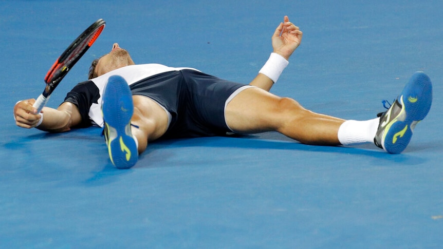 A tennis player holds his racquet while lying on the blue surface of a tennis court