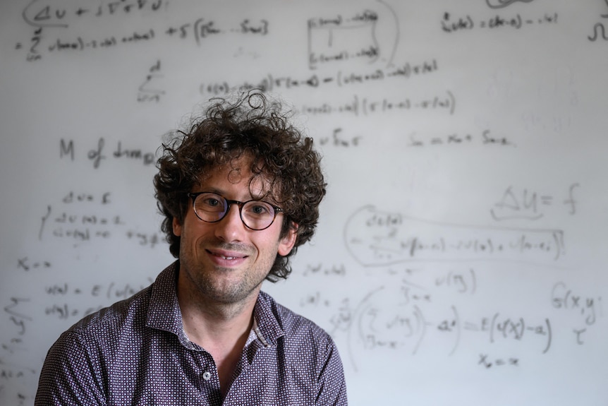 A man with curly hair and wearing glasses, stands in front of a white-board with maths equations