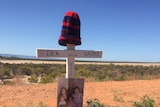 A cross with the words Dick and Naomi and a photo on it surrounded by flowers in bushland