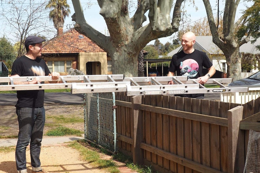 Two men stand either side of a fence while passing a ladder across