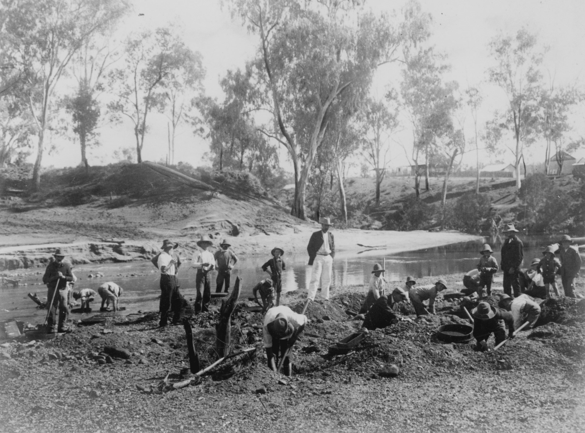 Black and white image of men in coats and pants crouching or standing at creek bed sieving for gems and holding pick axes.