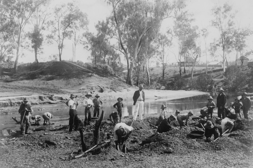 Black and white image of men in coats and pants crouching or standing at creek bed sieving for gems and holding pick axes.