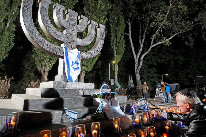 Menorah-shaped monument at Babi Yar memorial site, surrounded by candles and Jewish man praying.