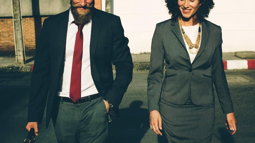 A man and woman in office attire walk towards the camera outside in sunshine