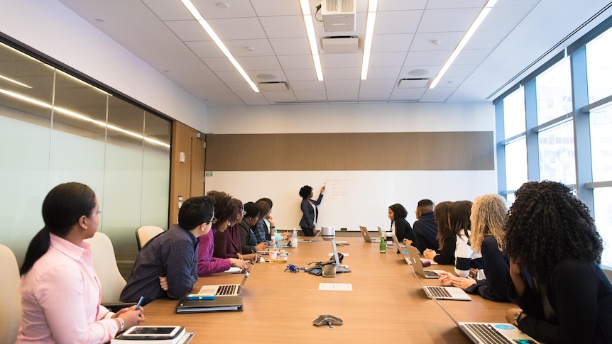 Conference room full of people of different nationalities looking down a table at a woman drawing on a whiteboard.