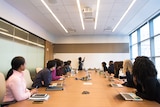 Conference room full of people of different nationalities looking down a table at a woman drawing on a whiteboard.