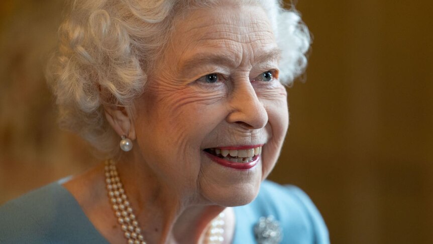 closse up image of queen elizabeth II smiling, wearing blue blouse with pearl earrings and necklace