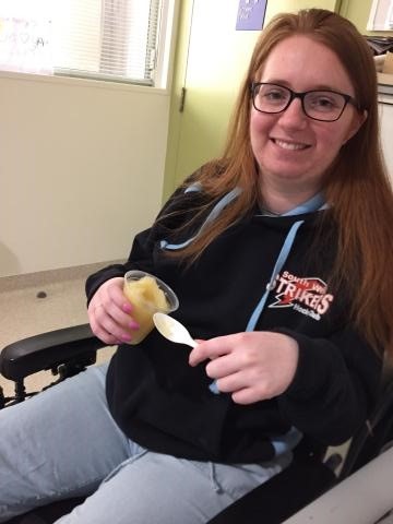 Jasmine is sitting, wearing a navy blue hoodie and holding a cup of apple puree and a spoon.