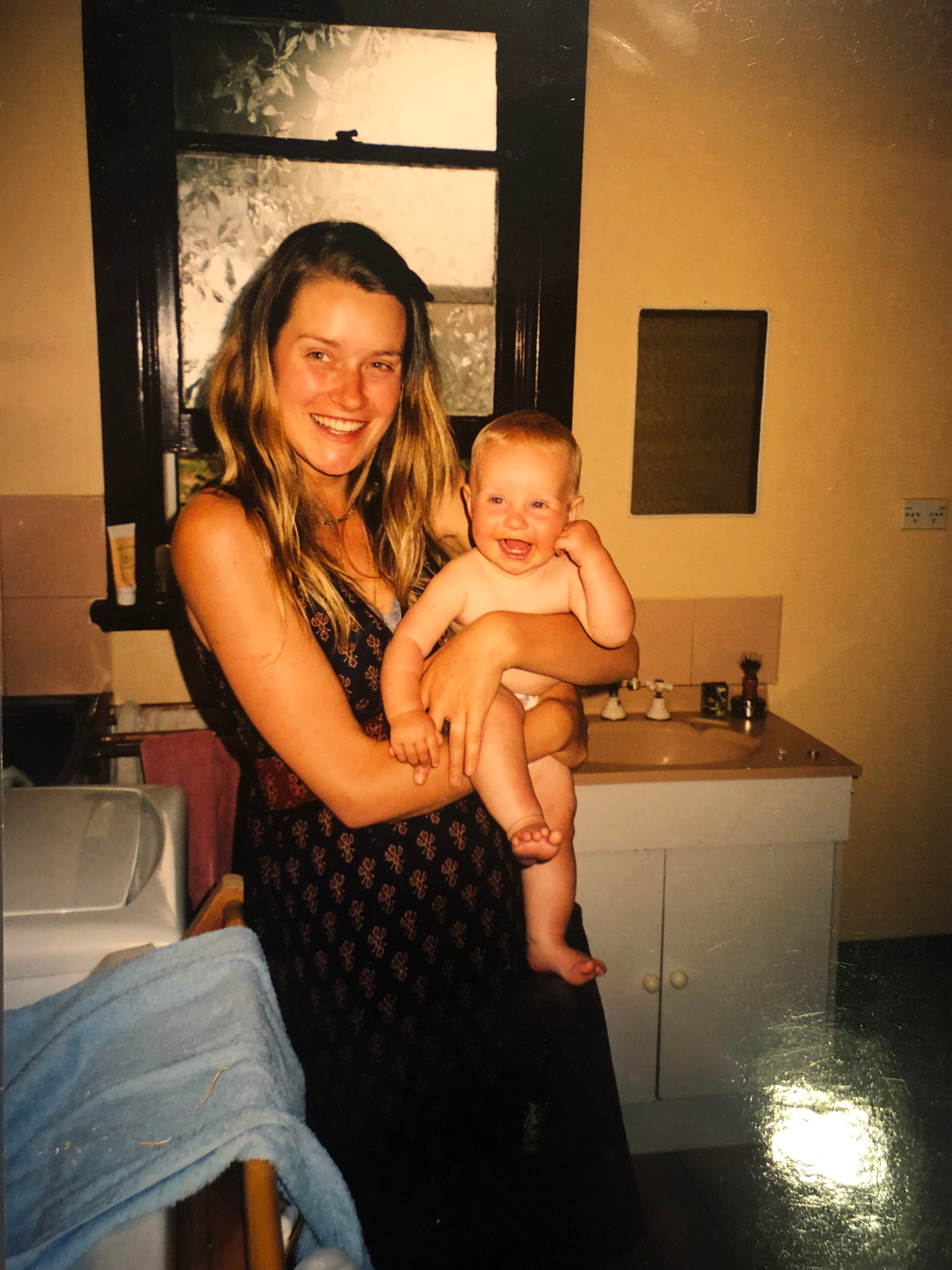 Blonde white woman in patterned black and red dress holds a smiling baby in a bathroom.