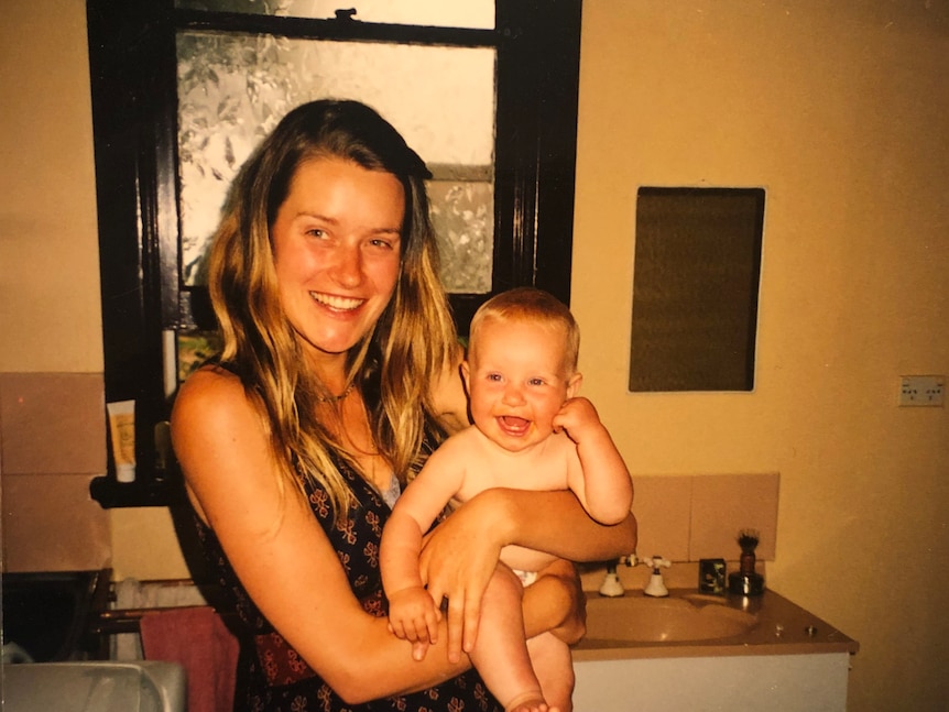 Blonde white woman in patterned black and red dress holds a smiling baby in a bathroom.
